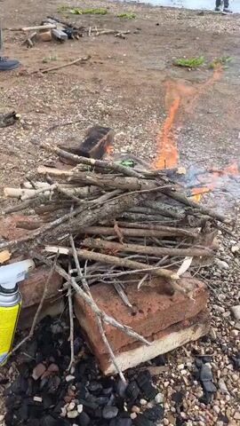A campfire is burning on a gravelly surface, with several sticks and logs piled together. Some of the sticks are burning intensely, with flames rising several feet into the air. In the background, a person's feet are visible, as well as some scattered debris. Two bricks are supporting the wood for the fire. There is smoke rising from the fire, and the overall scene suggests an outdoor activity.