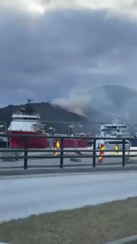 The video shows a hillside fire burning in a mountainous area, with a large cargo ship docked in the foreground. Smoke billows from the fire, obscuring parts of the mountainside. The fire seems to be traveling down the slope. In the foreground, there's a road or path with a metal railing. A large red and white cargo ship is visible on the water. Another smaller, white ship can be seen further away. The sky is overcast, and the overall lighting suggests it is either early morning or late afternoon.