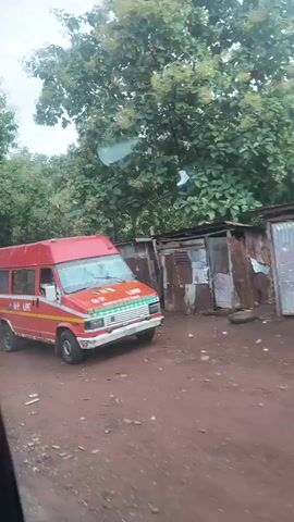 The video begins with a close-up of a red and blue bus with "G.P. LINE" written on its side, parked next to a simple wooden market stall. A woman stands by the open back door of the bus, and several people are visible inside the bus. The camera then pans to the left, showing more of the market stalls, which are made of wood and corrugated metal and appear to be selling produce. The stalls are set up along a dirt road, and lush green trees and foliage surround the area. The weather appears overcast. The bus moves forward slightly, and the camera continues to pan, revealing more stalls and people sitting and standing around them. A solar-powered street light is visible in the distance. The video ends with a wider shot of the market scene.