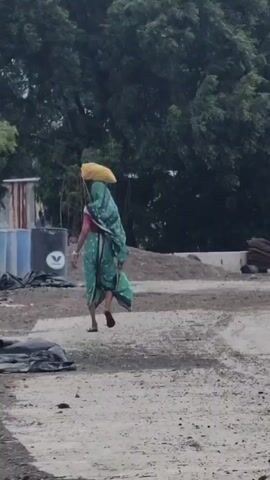 A person walks away from the camera, carrying a large yellow object on their head and a white plate in their right hand. The person is wearing a green and blue sari. The ground is dirt and gravel. In the background, there are trees, a corrugated metal structure, and some large blue and white tanks. The lighting suggests it is daytime, possibly overcast.