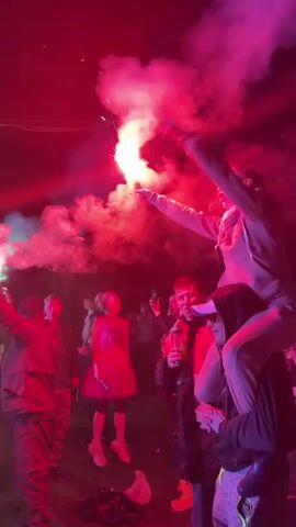 A night-time, low-angle, full shot from a concert shows a diverse crowd dancing and holding up flares that illuminate the scene with red and green light. In the foreground, a man with a beard holds a red flare and waves it above his head, creating a plume of smoke. Another man with a beard is visible in the middle ground, standing on the shoulders of another man while holding a red flare with sparks and smoke emanating from it. Behind them, people are dancing and cheering. The background shows a stage with performers and lights, surrounded by trees and a dark sky. The crowd is dressed in casual clothing, and some are wearing hats. The video is set to upbeat music, and the crowd appears to be enjoying themselves.