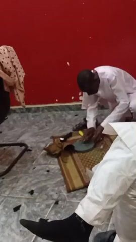The video shows two people in a room with a red wall. One person wearing a white robe and black socks is sitting on the floor, observing another person who is squatting and working on an object on a small prayer rug. The person squatting is also wearing a white robe and is using tools on what appears to be a disc-shaped object. There are various other items scattered on the floor, including what seems to be debris.