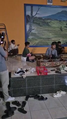 A video begins showing a group of people indoors, possibly practicing a musical performance. In the foreground, a person is filming with a camera. To the left, a young person is crouching, looking at a phone. In the center, another young person is sitting, holding a musical instrument, likely a gong or xylophone type instrument, and appears to be playing it. To the right, a young person is also sitting and appears to be playing a similar instrument, and is smoking a cigarette. Several other young people are visible in the background. The room has orange walls and a large mural of a landscape on the back wall. There are various items on the floor, including clothing, shoes, and musical instruments.