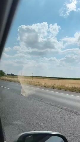 The media file captures a view from inside a vehicle, looking out the passenger side window. The scene depicts a road traveling through a rural landscape under a bright, partly cloudy sky. The road itself is asphalt with a white lane marker. On the right side of the road, there is a dry, grassy field bordered by a fence and some scattered bushes and trees. In the distance, a wind turbine is visible, suggesting an open, potentially agricultural area. The sun is shining, casting a glare on the window and creating a rainbow effect in one section. The overall impression is a sunny day, possibly late morning or early afternoon, suitable for travel. There is no visible text in the media file.