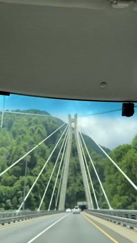 The video shows a view from inside a vehicle as it travels across a bridge. The bridge has tall concrete support towers with cables extending outwards. The surrounding environment is mountainous and covered in lush green trees. Several cars are visible on the bridge, moving in the same direction as the camera. The sky is blue with some clouds. The video is short, with timestamps indicating it lasts only a few seconds.