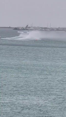The video shows a seaplane maneuvering on a body of water. The seaplane is painted in red and white, with the registry "8Q-MAT" visible on its tail. As the plane moves, it creates a spray of water behind it. In the background, there is a shoreline with various buildings and structures, indicating an urban or developed area. An orange buoy can also be seen in the water near the plane. The sky is overcast, suggesting a cloudy or rainy day.