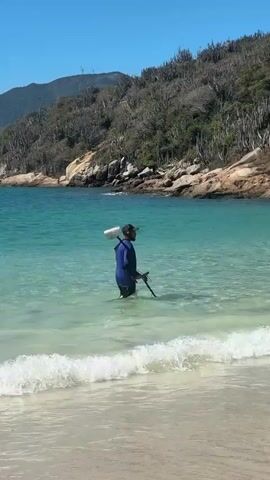 A person wearing a blue long-sleeved shirt and dark shorts stands in clear, shallow ocean water near a sandy beach. They are holding a metal detector with a black handle and circular sensor, and a shovel with a flat metal scoop. The person appears to be metal detecting in the water. In the background, there is a rocky shoreline with sparse vegetation and trees, leading up to a hillside under a bright blue sky. Gentle waves are breaking on the shore in the foreground.