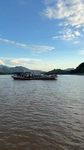 A wide shot captures a calm, muddy river under a bright blue sky filled with scattered white clouds. In the center of the frame, a long, flat-bottomed ferryboat travels across the water. The ferry is crowded with people standing and sitting, and appears to be carrying vehicles as well, though these are not clearly visible. The ferry has a red hull and a white superstructure. On the right side of the frame, a lush green, tree-covered hillside slopes down to the water's edge, with several other smaller boats docked along the shoreline. On the left side of the frame, distant mountains are visible. The scene suggests a tranquil, possibly rural setting, with the ferry serving as a mode of transportation for the people on board.