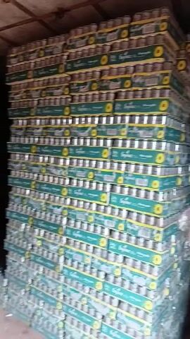 The video shows a storage area, possibly a warehouse or a large storeroom, filled with stacks of canned pineapple juice. The cans are packaged in cardboard boxes, with the brand name "Fayrouz" and "Pineapple" printed on them. The boxes appear to be stacked high against a wall, creating a large, dense structure of products. The lighting in the room is dim, making the details a bit difficult to discern.  Some loose boxes of the same product are on the floor to the left of the stack. There does not appear to be anyone visible in the video.