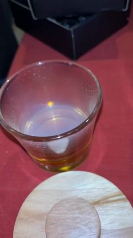 A medium shot shows a glass on a red surface containing a small amount of brown liquid. There is condensation on the glass. Next to the glass is a two-piece wooden lid. In the background is a black box with black circles and a bottle of Jack Daniels Tennessee Whiskey. A butane torch is next to the Jack Daniels bottle. There is music playing in the background.