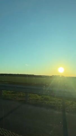The video shows a landscape view of a field and the sun setting in the distance, observed from inside a moving vehicle. The sky is a gradient of blue at the top, transitioning to orange and yellow near the horizon where the sun is situated. The sun appears as a bright, circular light source.

In the middle ground, there's a field with green grass, bordered by a tree line in the distance. A fence can be seen running along the perimeter of the field. In the immediate foreground, a grey guardrail runs horizontally across the bottom of the frame, partially obscuring the view of the field. 

The camera is positioned within a moving vehicle, as evidenced by the slight blurriness of the passing landscape and the visible interior of the vehicle at the very bottom of the frame.