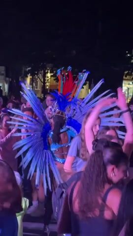 A person in a vibrant blue and red feathered costume with a headdress is dancing. They are surrounded by a crowd of people who are also moving and appear to be enjoying the performance. The lighting is dim, suggesting it is nighttime or an indoor venue with stage lighting. The crowd is mostly obscured by the central figure and other audience members. The costumes and general atmosphere suggest a festival or celebration.