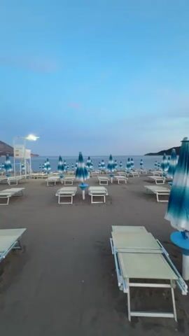 The video shows an empty beach at dusk or dawn. Numerous beach chairs and umbrellas are arranged in rows on the sand. The sky is a clear, pale blue with a few wispy clouds. The ocean is calm and stretches out to the horizon. In the background, a distant coastline with buildings is visible. There is a lifeguard stand on the left side of the frame, also empty. The lighting suggests it is either early morning or late evening.