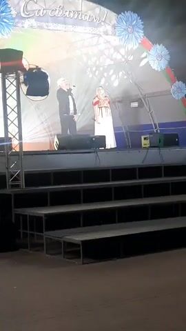 TWO PEOPLE STAND ON A STAGE. A MAN ON THE LEFT WEARS A DARK JACKET, LIGHT PANTS, AND A WHITE HAT. HE HOLDS A MICROPHONE IN HIS RIGHT HAND AND APPEARS TO BE SPEAKING OR SINGING. A WOMAN ON THE RIGHT WEARS A WHITE DRESS WITH EMBROIDERY. SHE HOLDS A MICROPHONE IN HER LEFT HAND. THE STAGE IS ILLUMINATED WITH STAGE LIGHTS. BEHIND THE PERFORMERS, A LARGE BANNER WITH FLORAL DECORATIONS IS VISIBLE. THE STAGE IS RAISED ABOVE A SERIES OF STEPS OR BLEACHERS. THE ENVIRONMENT APPEARS TO BE OUTDOORS, POSSIBLY AT AN EVENT OR FESTIVAL.