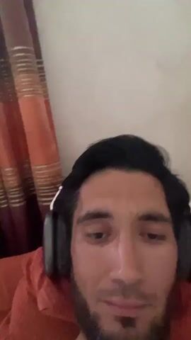 The video shows a close-up, slightly downward-angled selfie of a man with dark hair and a beard, wearing black headphones. He is lying down, as indicated by the background, which includes an orange-tinted curtain with vertical stripes and a red blanket or pillow. The man blinks and smiles at the camera.