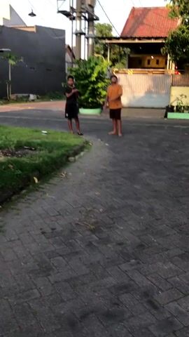 Two young males are standing on a paved driveway outdoors. The male on the left is wearing a black t-shirt and black shorts. He is holding his hands up as if to catch something. The male on the right is wearing an orange t-shirt and brown shorts. He appears to be throwing something upwards. There is a grassy area to the left of the driveway and a building with a red roof in the background. A utility pole with wires is visible behind the building. The lighting suggests it is daytime.