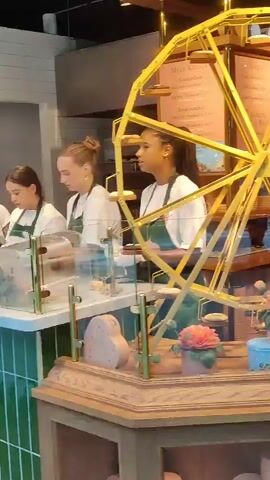 This video shows the interior of a bakery or dessert shop. In the foreground, there is a wooden display counter topped with a glass case. Inside the glass case are decorative items, including a gold-colored Ferris wheel structure with small pastries or cookies displayed on its spokes, and several colorful artificial flowers in blue and pink pots. Behind the glass case, two young women wearing green aprons over white shirts are working behind a counter. One woman, with dark curly hair pulled back in a ponytail, is standing behind the Ferris wheel display, facing to her left and appears to be preparing something. To the left of her, another woman with blonde hair in a bun is also wearing a green apron and white shirt, and she is facing to her left, looking towards the display. Further back, behind the counter, there are shelves with baked goods, and another woman is visible, also wearing an apron. The background includes menus displayed on the wall and shelving units filled with more baked goods. The lighting is bright, suggesting an indoor commercial setting.
