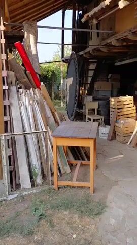 The video opens with a view of a cluttered outdoor area. Piles of wood, including planks, beams, and ladders, are stacked against a wooden structure. A small orange table with a dark top and a chair are positioned in the foreground. The camera then pans to a different section of the yard, revealing a grassy area with several wooden posts and what appears to be a large vine-covered structure overhead. Further on, a white van is partially visible on the left side of the frame. The scene suggests a backyard or storage area with various materials and vehicles present. The environment appears to be a garden or yard with trees and vegetation.