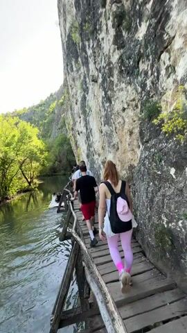 A group of people are walking single file along a wooden boardwalk trail constructed along a river. To the right of the trail is a tall rock wall, and to the left is the river. The water is calm with a slight ripple. Trees with green leaves line the opposite shore. The sky is bright. The boardwalk is made of wooden planks and has a rough wooden railing along the river side. The person closest to the camera is a red-haired woman wearing a white tank top, a white skirt, bright pink leggings and grey athletic shoes. She has a black backpack with a small pink hat attached. She pauses and puts her hands up to her hair. In front of her are two men, one wearing a black t-shirt and red shorts, and the other wearing a white shirt. Other people are farther ahead on the boardwalk. A small boat is visible on the river.