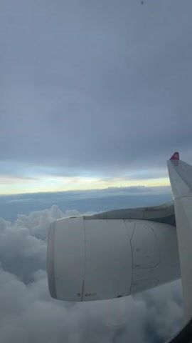 From an airplane window, the view includes a white plane engine and a portion of the wing with the Turkish Airlines logo (a red bird). Below, fluffy white clouds fill the sky, with a thin band of yellowish light peeking through on the horizon, suggesting the presence of the sun behind the clouds. Above, the sky is covered with overcast, grey clouds. The scene is still and provides a sense of flight above the clouds.