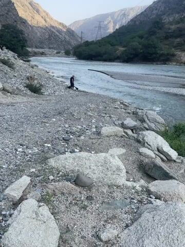 Two individuals are standing near the edge of a wide, fast-flowing river. The riverbed appears rocky with areas of shallow water and deeper currents. The surrounding environment is mountainous and arid, with steep, barren slopes on either side of the river. Vegetation is sparse, consisting mainly of scrubby bushes and small trees along the riverbanks and lower mountain slopes. In the distance, power lines and towers are visible on the mountainside, indicating human infrastructure in the area. The ground in the foreground is covered with stones, pebbles, and larger rocks. One individual is partially obscured by the other, and they appear to be looking out at the river. The overall impression is one of a remote, natural landscape.