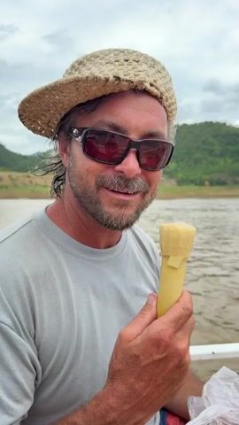 A man wearing a straw hat and sunglasses on a boat takes a bite out of a yellow ice cream cone. He is holding the cone in his right hand and has it in his mouth. The background shows a body of water and green hills.