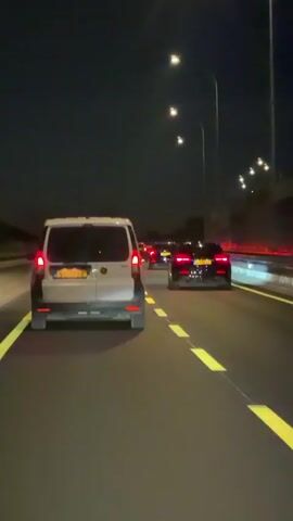 A first-person perspective video shows a convoy of vehicles driving on a highway at night. The road is dark, illuminated by streetlights and the headlights and taillights of the vehicles. The camera is positioned inside the rear of a white van, facing backward towards the road. A black sedan is visible behind the white van. Further ahead, other vehicles can be seen with their taillights illuminated. Yellow dashed lines mark the lanes on the asphalt road. The environment appears to be an open highway with streetlights lining the side.