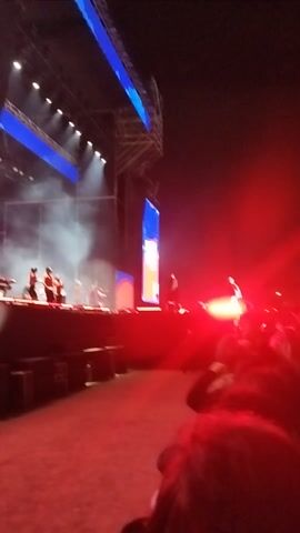 This video captures a vibrant outdoor concert or festival at night, set in Maipú, Chile.

The scene opens with a dynamic view of a large stage featuring multiple performers, possibly dancers, partially enveloped in stage smoke. Bright white spotlights illuminate the scene, flanked by prominent blue LED screens on the left and a red screen on the right. The stage structure is elaborate, with scaffolding and various lighting fixtures.

The camera swiftly pans to a densely packed audience, primarily lit by a strong red hue from stage lighting. Numerous attendees are holding up their mobile phones, many with active screens or flashlights, adding to the energetic visual display. The crowd appears engaged and enthusiastic, swaying to the music.

Loud music and enthusiastic cheering are audible throughout the clip, indicating a lively atmosphere. The time of day is clearly night, as the sky is dark and all illumination comes from artificial sources. No discernible text is visible. The video is shot handheld and has some motion blur due to quick panning and low light.