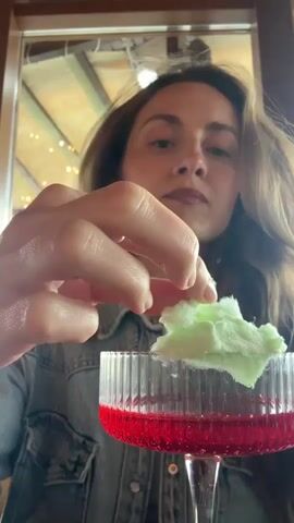 A woman is holding a piece of light green cotton candy in her right hand. The cotton candy is near the top of a clear stemmed glass, which contains a red liquid. The woman puts a small piece of the cotton candy on her tongue. She looks up and chews. She looks back toward the camera and smiles. The woman is wearing a denim jacket over a white shirt. Her brown hair is parted to her left. The background contains a window with natural light. The top of a tall, brown, wooden frame of some kind is visible, as well as a light fixture with multiple light bulbs.