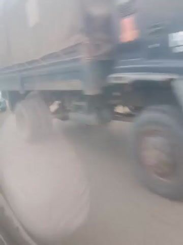 This video captures a street scene from a low angle, possibly from inside a vehicle. The image is blurry and tilted, making it difficult to discern many details. However, the footage shows several vehicles, including a white van and what appears to be a smaller light-colored car. There are also glimpses of what might be motorcycles and bicycles. People are visible, but their features are indistinct due to the blurriness and distance. The environment appears to be a street with some trees and buildings in the background. The overall impression is of a busy, perhaps congested, area.