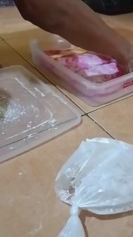 The video shows hands working with a pink and white marbled dough-like substance in a clear plastic container. Another clear plastic container with some white powder is visible. A plastic bag containing white powder is also in the foreground. The setting appears to be a floor with tiled surfaces. The hands are seen manipulating the dough, possibly preparing it for some use.