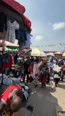 The video shows a crowded outdoor market scene with many people walking around and looking at clothes and other goods. The market stalls are covered with colorful umbrellas and awnings. Clothes are hung up and piled on tables, and people are browsing through them. In the foreground, a man in a red and black jersey is looking at clothes on the ground. In the middle ground, a woman wearing a light brown shirt and blue jeans walks towards the camera, stepping down some steps. The overall environment appears to be busy and lively. The sky is partly cloudy and there are strings of pennants overhead.