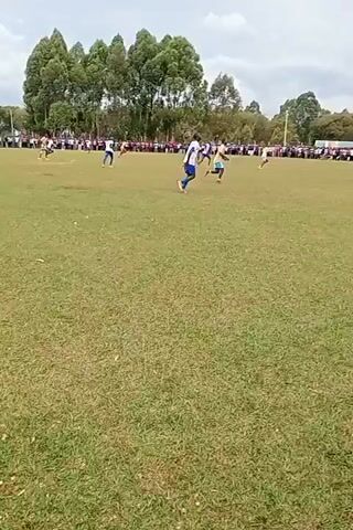 The video shows a soccer game being played on a grassy field. Spectators line the perimeter of the field, watching the game. A player wearing a blue and white jersey is running towards the goal. Another player is close behind him. In the foreground, a man wearing a white jacket with "GOR MAHIA FC" printed on the back is also watching the game. The sky is overcast.