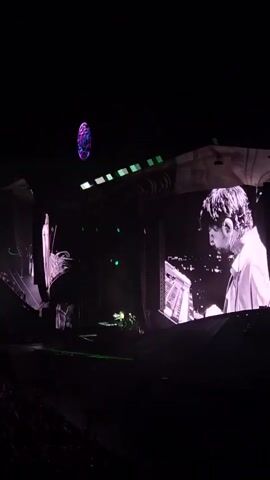 A wide shot shows a concert stage at night. The stage is dark with a large screen displaying a black and white image of a person's face. The person appears to be a man wearing a suit and hat. Green laser lights are visible on the stage. In the foreground, a crowd of people can be seen watching the performance. The ceiling above the stage has a circular projection with purple light.