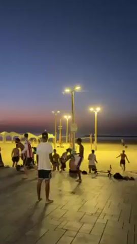 The video shows a group of young people playing a game with a ball on a paved area next to a beach at dusk. There are at least ten people visible, some shirtless and others wearing shorts and t-shirts. They are passing and kicking a ball among themselves, illuminated by several lampposts that cast a warm light. The beach is in the background, with other people on the sand and some tents or canopies visible in the distance. The sky is a mix of dark blue and the orange glow of the setting sun. People walk by in the foreground, seemingly uninvolved with the game.