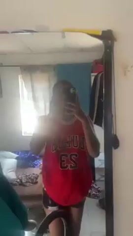 A person wearing a red Chicago Bulls jersey and black and white patterned shorts stands in front of a mirror, holding a smartphone and taking a selfie. The mirror is rectangular and framed in black. The room behind the person is somewhat cluttered. There is a window to the left of the mirror with a translucent curtain partially drawn. A bed covered in clothing and blankets is visible. A green garment hangs on the left side of the frame. Clothes hang in the background to the right of the mirror. The room appears to be brightly lit by natural light.