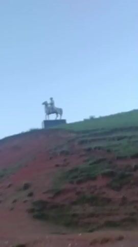 A blurry video shows a statue of a person on a horse silhouetted against a pale blue sky. The statue is positioned on a reddish-brown hill with some sparse green vegetation. As the video progresses, the statue appears to move downhill, with the rider's arm extended. The moon is visible in the sky in the first few seconds of the video. The overall quality of the video is poor and pixelated.