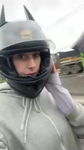A person wearing a gray hoodie and a black motorcycle helmet with two ears on top is riding on the back of a motorcycle. The person is looking at the camera, and the person driving the motorcycle is wearing a green and black helmet. The background is blurry and appears to be a highway. The weather appears overcast.