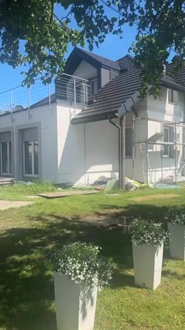 The video pans across the side of a house that appears to be undergoing renovation. Scaffolding is erected against the white exterior walls. There are several windows visible, and a balcony with a metal railing on the upper floor. In the foreground, a well-maintained lawn is visible, with decorative planters containing white flowers. The scene is lit by natural sunlight, suggesting it is daytime.