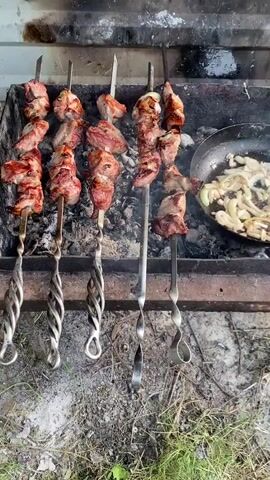 A close-up overhead shot shows several skewers of meat being cooked over coals. The meat is chunks of pork, some with fat, and appears to be browning and slightly charred. The skewers are made of metal with decorative twisted handles. To the right, a black frying pan contains sliced onions being cooked. Smoke rises from the coals and the cooking food. The scene is outdoors, with dirt and dry grass visible in the foreground, and what appears to be a rustic metal grill structure.