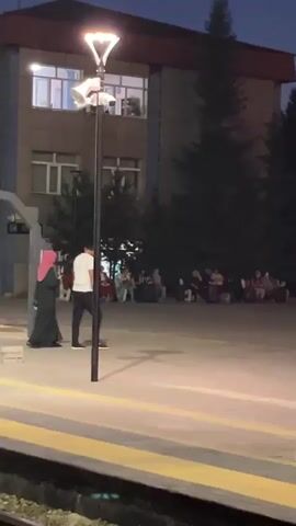 The video shows an outdoor scene at dusk or night. In the foreground, a yellow line marks the edge of a platform, possibly a train station. To the right, a large, cylindrical concrete pillar with a white base and two yellow warning signs is prominent. Beyond this, an area with people sitting on benches and on the ground is visible, with trees and buildings in the background. To the left, a street lamp with a white shade illuminates the area. Further back, a temporary structure resembling scaffolding with banners showing pictures of people is erected. Several individuals are visible in the scene, some walking and others seated. The lighting suggests it is evening or night, with artificial lights providing illumination.