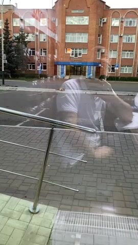 The video shows a person in a white shirt lying face down on the ground in front of a brick building. The person's arm is extended, and they appear to be writing on a piece of paper. There are reflections in a window in the foreground, which obscure some of the view. The environment appears to be an urban setting, with a street and sidewalk visible. There is a bus stop in the background. The person's head is partially obscured by the reflections. The video appears to be taken from inside a building, looking out. The person is lying on a paved surface. There are some objects on the ground near the person, including a plastic bag and a bowl.