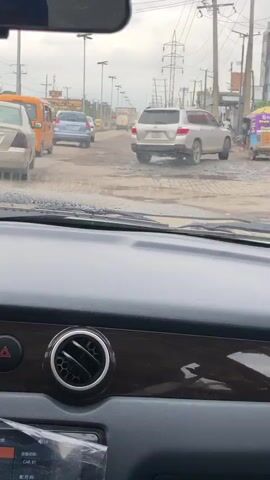 The video begins inside a car, looking out through the windshield at a busy street. The road is in poor condition, with many potholes and puddles. Several vehicles are visible, including cars, a yellow bus, and motorcycles. On the right side of the road, there are buildings, street vendors with umbrellas, and a sign that says "POS". People are walking and standing on the side of the road. The sky is overcast. The camera is relatively steady, suggesting the car is moving slowly. The interior of the car is visible in the foreground, with a black dashboard and a wooden trim piece.