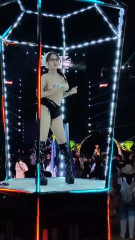 A woman is dancing on a small platform inside a decorative metal structure covered in small lights. She is wearing a bikini top, black shorts, and tall black platform boots. She is also wearing a white bandana on her head and glasses. She is smiling as she dances. The platform is lit up from underneath, and bright white stage lights are shining in the background. A crowd of people can be seen watching her in the background, some holding up their phones to take photos or videos.