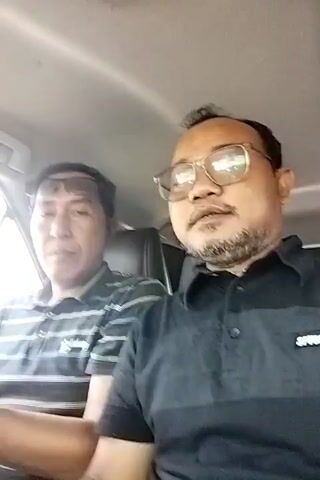 The video shows two men sitting in the front seats of a car. The man in the foreground, wearing glasses and a black polo shirt with horizontal stripes, is looking directly at the camera and speaking. The man to his left, wearing a striped polo shirt, is also looking at the camera, but appears to be listening or waiting to speak. The environment appears to be the interior of a car, with sunlight coming through the windows. The man in the foreground gestures with his hand as he speaks. The man in the background has sunglasses perched on his forehead.