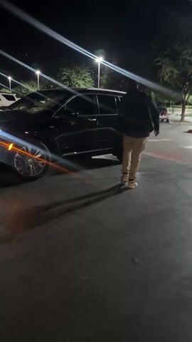 At night, a man dressed in a black hoodie, tan pants, and sneakers stands near a black SUV in a parking lot. The rear passenger door of the SUV is open. Streetlights cast long shadows on the asphalt, and trees are visible in the background. The man appears to be talking with someone, then he shuts the car door.