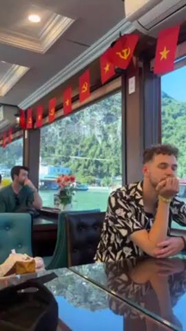 The camera shows a man sitting at a table on a boat. He is wearing a black-and-white patterned shirt and has his hand on his chin. The boat is decorated with Vietnamese flags. Through the window, one can see a bay and lush, green islands. Another man sits at a nearby table. A backpack and other personal items sit on the table. An air conditioner is mounted on the ceiling.