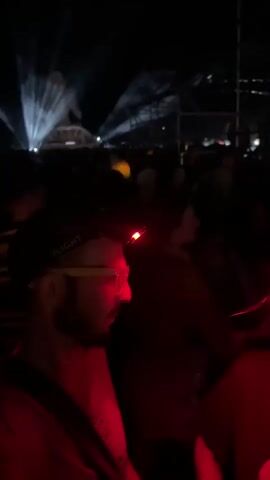 The video begins with a close-up of a man wearing glasses and a headlamp with a red light. He is in profile, looking to the left, and appears to be outdoors at night. The background is dark, with some blurry lights and figures. The camera then pans, and the man's headlamp illuminates the scene. The view becomes shaky, suggesting movement, and the red light dominates the frame, creating streaks and reflections. The scene briefly shows blurry lights that appear to be from a moving vehicle or a distant event. The video then transitions to a wider shot of a dark outdoor space with a large, illuminated sculpture of a seated figure that appears to be made of light or ice, emitting white and blue beams of light. A crowd of people is gathered in front of the sculpture, facing it. The lighting suggests a performance or event taking place at night.