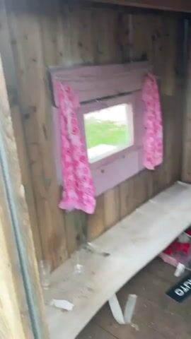 The video shows the interior of a wooden structure, possibly a playhouse or shed. The walls are made of rough-hewn wood planks. There is a window with pink trim and a pink patterned curtain. A long, light-colored wooden bench runs along one wall. On the bench, there are a few small clear plastic cups and some white debris. To the right of the bench, there are some colorful objects, possibly toys or supplies, in plastic bags.

In the center of the frame, a young girl with brown hair and bangs is visible. She is wearing a purple t-shirt with a cartoon character on it and black shorts. She is looking at the camera and appears to be standing or sitting. Her right hand is raised, touching her shoulder.

A green plastic ride-on toy with pink wheels is visible in the right side of the frame. The floor is also made of wooden planks and appears somewhat dusty. The overall impression is of a rustic, possibly abandoned or seldom-used space.