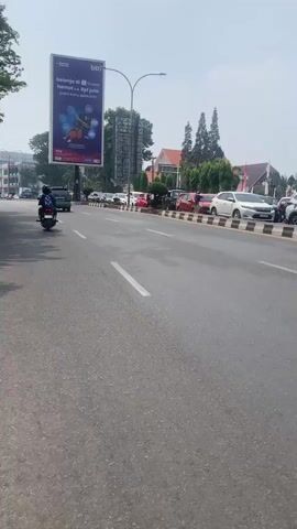 The video captures a street scene from the perspective of a person on a motorcycle or scooter. The camera is facing forward, looking down the road. On the left side of the road, there is a large billboard advertising for "BTN" and "Shopee" with text in Indonesian. The billboard features images related to shopping and banking. Further down the road on the left, there are several cars parked and some buildings. On the right side of the road, there are more cars driving in the same direction as the camera. A black minivan is visible in the right lane. The road is paved and appears to be a multi-lane street with white lane markings. The sky is visible in the background and appears to be clear and bright, suggesting it is daytime. Trees and some buildings are also visible in the distance. The primary action is the movement of the camera vehicle and other vehicles on the road. A person on a scooter in the foreground is the most prominent individual, seen from the back, wearing a black jacket with a logo and a helmet. Another person on a motorcycle is also visible in the distance on the left. The overall environment seems to be an urban or suburban street with moderate traffic.